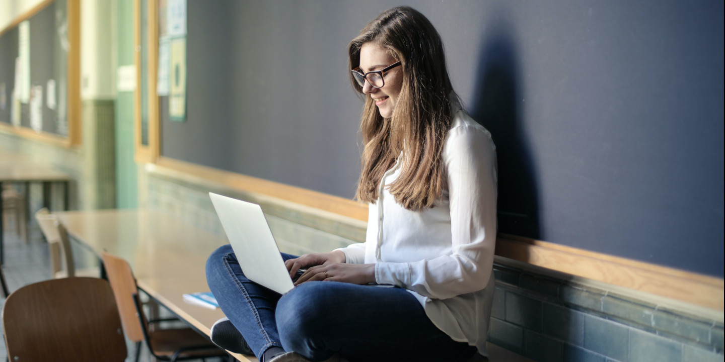 Eine junge Frau sitzt im Schneidersitz auf einem Tisch, der vor einer Tafel steht und schau in ihren Laptop.