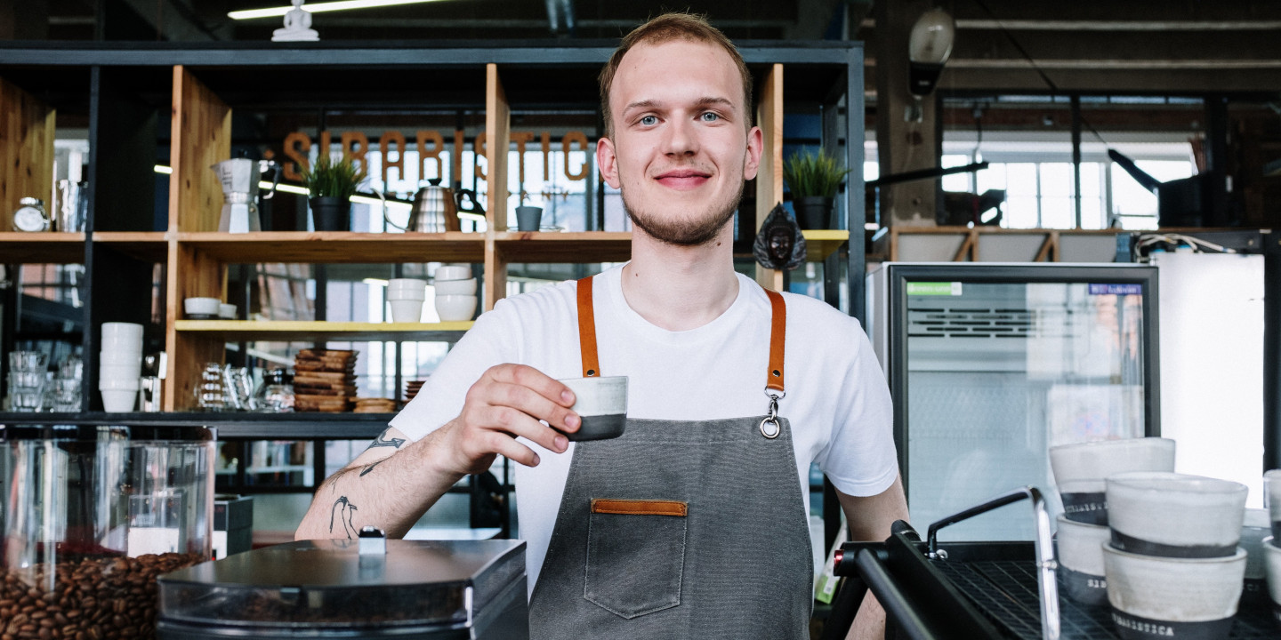 Ein junger Mann mit Schürze und einem Kaffee in der Hand steht in einem Cafe und lächelt in die Kamera.